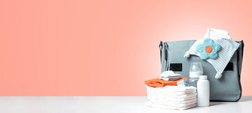 A neatly arranged baby diaper bag with essential items, including diapers, baby wipes, a baby bottle, a pacifier, and a soft cloth with a blue flower decoration, set against a pastel pink background.