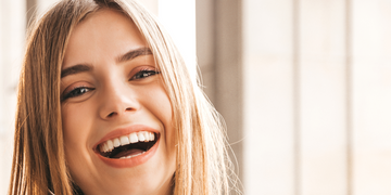 A close-up of a young woman with long, straight blonde hair laughing joyfully. Her bright smile and white teeth are highlighted, with soft natural light coming through large windows in the background.
