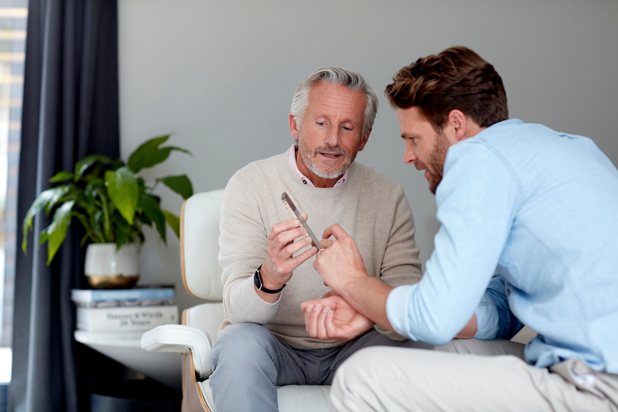 An older man wearing an Aktiia 24/7 Blood Pressure Monitor on his wrist demonstrates its use to a younger man in a home setting. They are engaged in conversation, discussing the device's features.