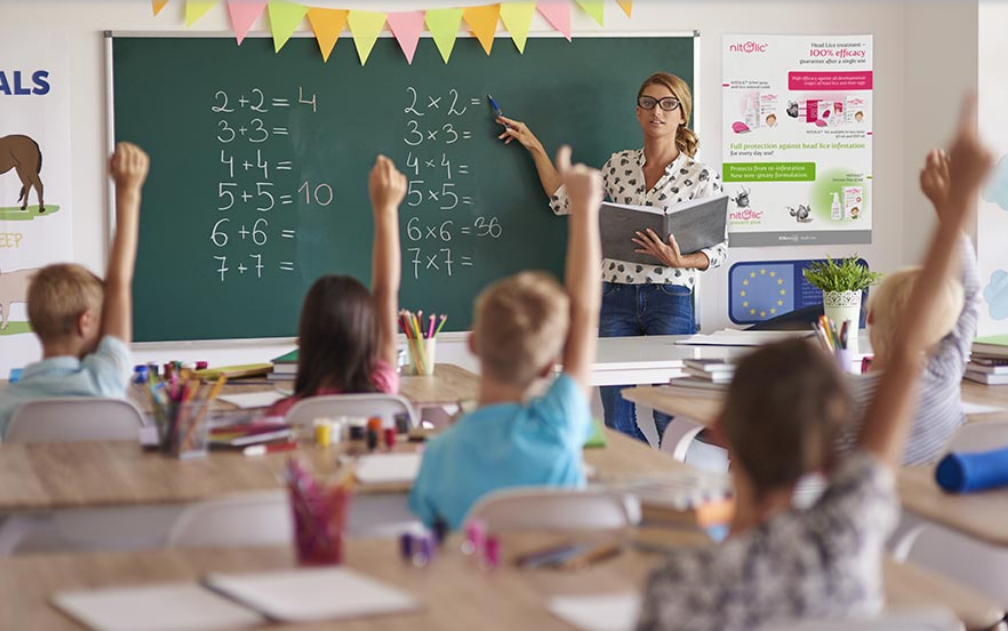 A classroom scene featuring a teacher in glasses and a white blouse with black polka dots standing at the front, writing on a chalkboard filled with math equations. Several young students sit at wooden desks, raising their hands enthusiastically. The classroom is decorated with colorful banners, educational posters, and a visible advertisement for a head lice treatment product on the wall. Natural light fills the room, creating a bright and engaging learning environment.