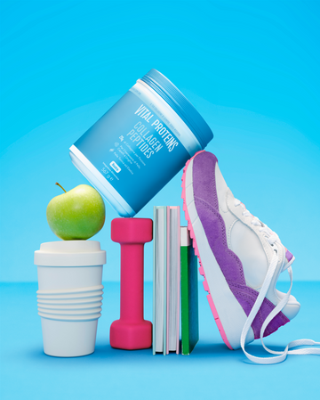 Vital Proteins Collagen Peptides container balanced on a stack of wellness items, including a green apple, coffee cup, pink dumbbell, books, and a purple sneaker, against a bright blue background.