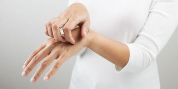 A person wearing a white long-sleeve shirt scratching the back of their hand, possibly indicating skin irritation, dryness, or an allergic reaction. The background is neutral grey.