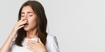 A young woman with long brown hair wearing a white t-shirt appears to be sneezing or having difficulty breathing. She is pinching her nose and touching her chest, with an expression of discomfort against a neutral grey background.