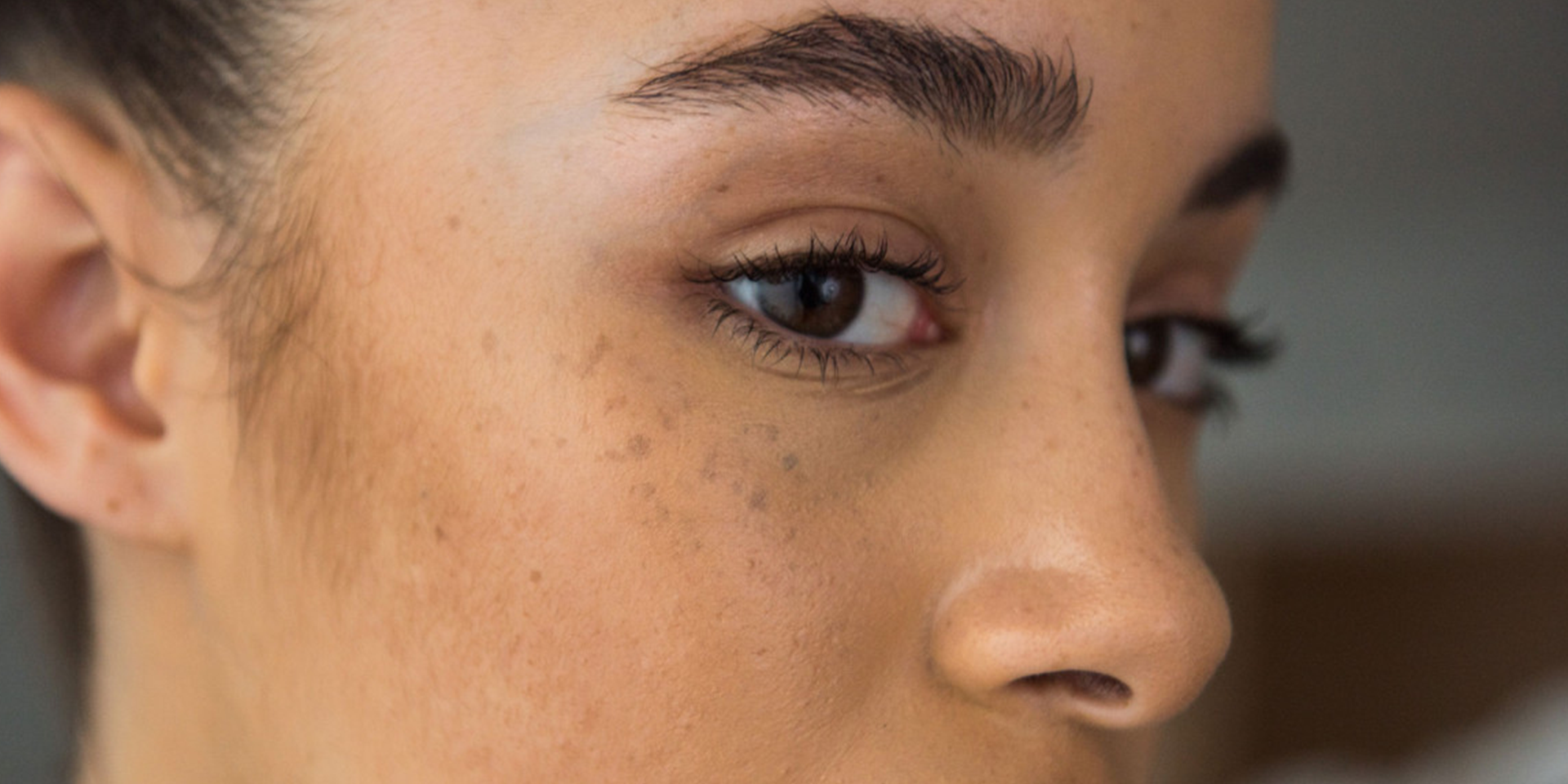 A close-up of a young woman's face, highlighting her brown eyes, thick eyebrows, and natural freckles. The soft lighting emphasizes her smooth skin and subtle imperfections.