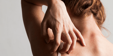 A close-up of a woman with light skin and brown hair scratching her upper back. Her posture and hand movement suggest discomfort or itchiness, with a neutral grey background.