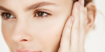 "A close-up of a young woman with smooth, radiant skin, gently touching her face. Her brown eyes, well-groomed eyebrows, and natural glow are highlighted against a soft, neutral background.