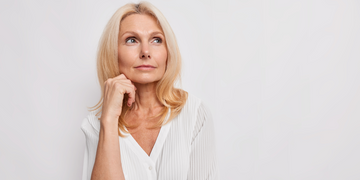 A mature woman with blonde hair and a thoughtful expression, resting her chin on her hand. She is wearing a white blouse and looking slightly upward against a plain white background.