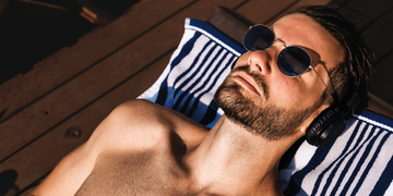 A bearded man wearing sunglasses and headphones, relaxing on a lounge chair with a blue and white striped towel, sunbathing on a wooden deck.