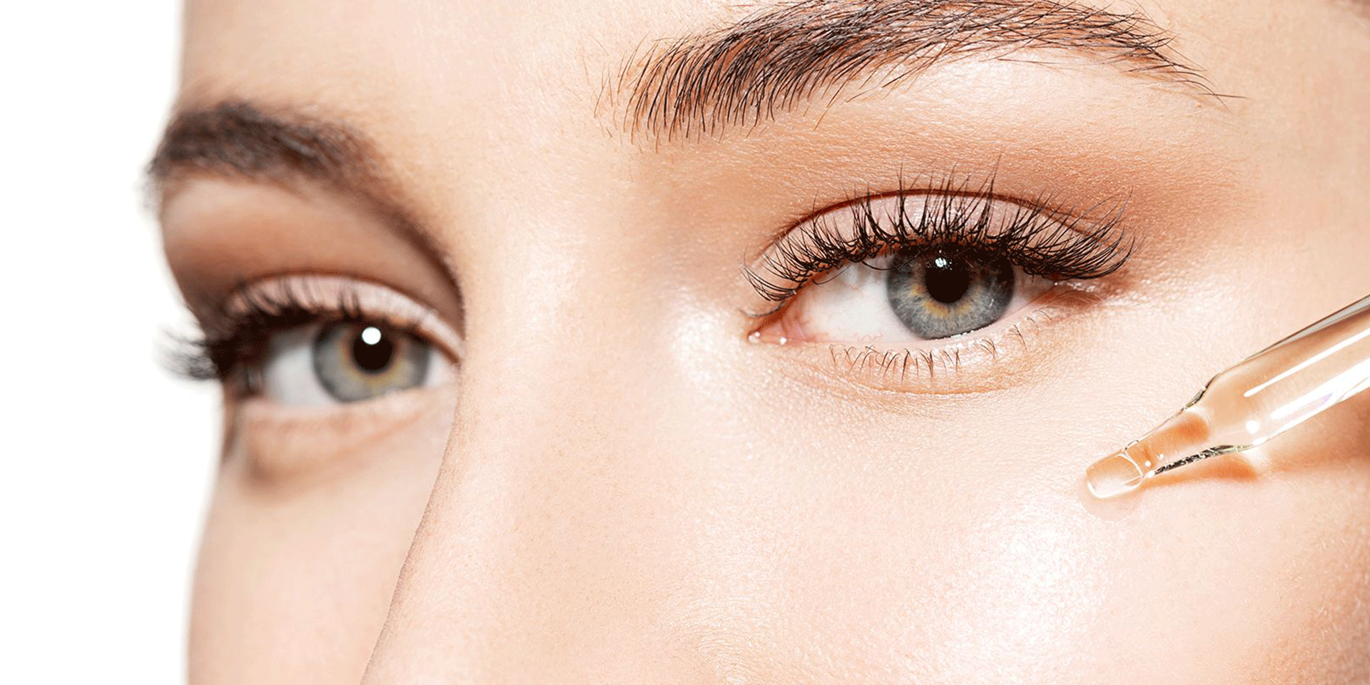 A close-up of a woman's face with smooth, glowing skin as she applies a skincare serum under her eye using a dropper. Her well-groomed eyebrows, long eyelashes, and light-coloured eyes are in focus.