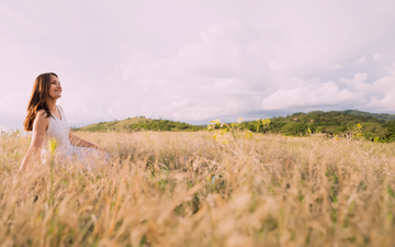 A woman in a white dress sitting in a golden field of tall grass, smiling as she enjoys the serene landscape. The background features rolling green hills and a cloudy sky, creating a peaceful and natural atmosphere.