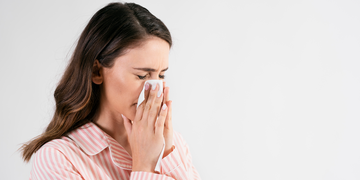 A young woman with long brown hair wearing a pink striped pajama top, holding a tissue to her nose while appearing to sneeze or have a cold. She has a pained expression against a plain white background.