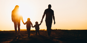 A silhouette of a family of four—two parents and two children—holding hands while walking outdoors at sunset. The warm sunlight creates a scenic and heart warming atmosphere.