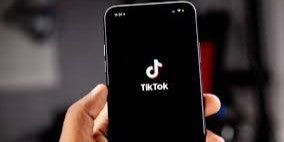A person holding a smartphone displaying the TikTok logo on a black screen. The background is blurred, suggesting an indoor setting.