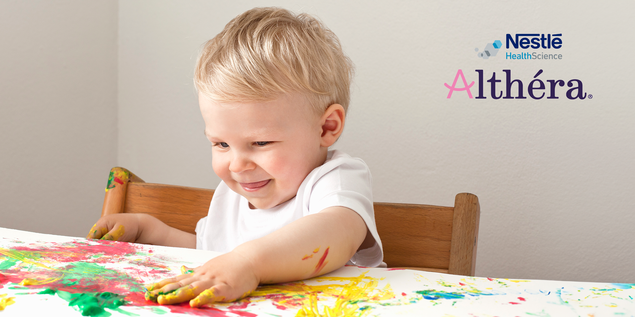 A cheerful toddler with blonde hair is finger-painting with vibrant colors on a white surface, smiling with delight. The Nestlé Health Science and Althéra logos are displayed in the top right corner. Althéra is a specialized infant formula for managing cow’s milk protein allergy, available at shop.prohealth.com.mt and pharmacies across Malta & Gozo.