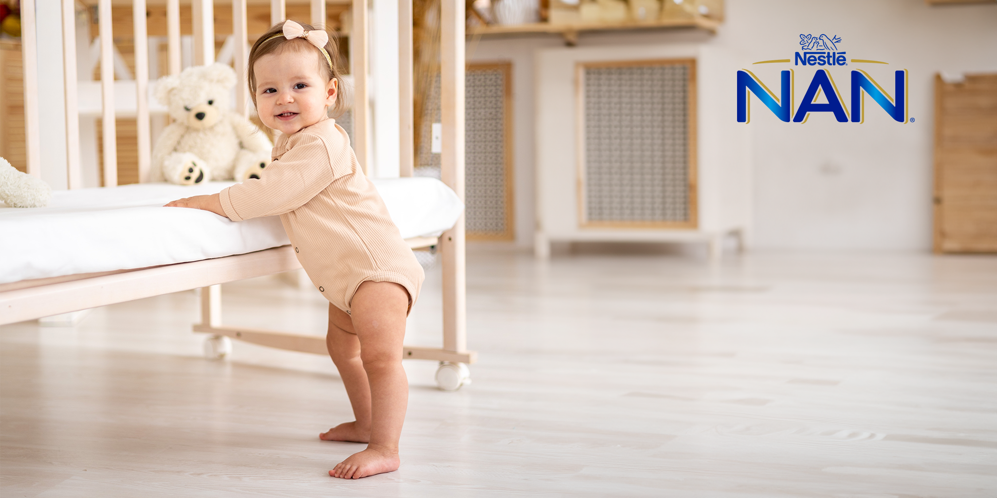 A Nestlé NAN advertisement featuring a happy baby girl standing and holding onto a wooden crib in a beautifully designed nursery. She is dressed in a beige onesie with a matching headband and has a joyful expression. A soft teddy bear sits on the crib, adding warmth to the cozy setting. The background is bright and airy, with modern wooden furniture. The Nestlé NAN logo is positioned in the top right corner, highlighting the brand’s infant formula products.