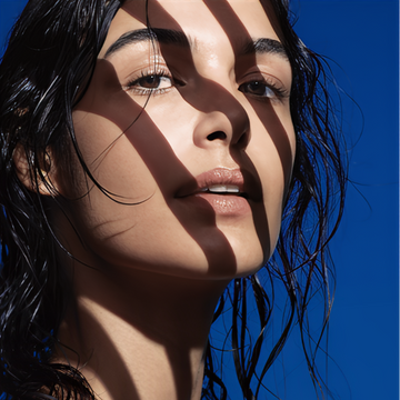 Close-up of a woman with wet hair against a vivid blue background, her face partially covered by bold shadows creating striped patterns. Her clear skin and natural makeup are highlighted by the sunlight.